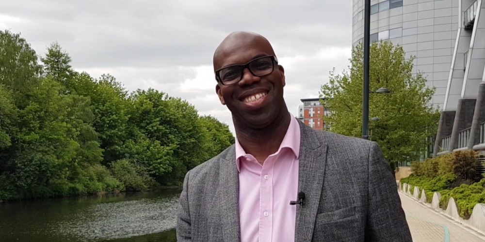 A smiling Cleveland Henry in a suit next to a river with modern office buildings in the background