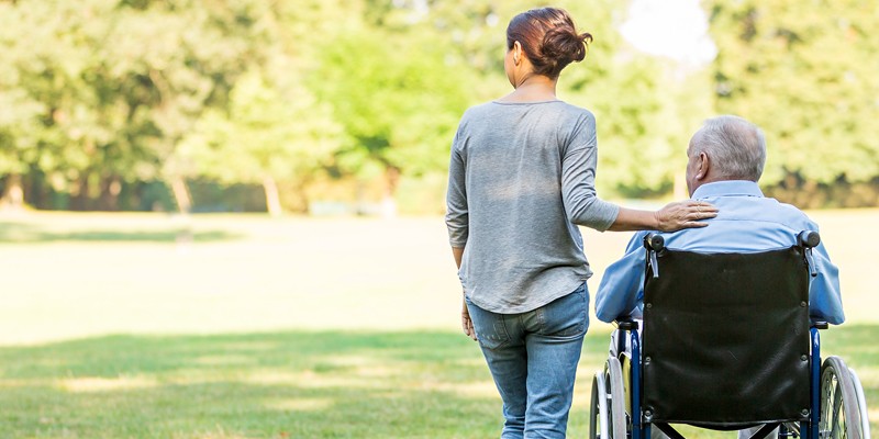 Younger woman puts arm around elderly man in wheelchair photographed from behind