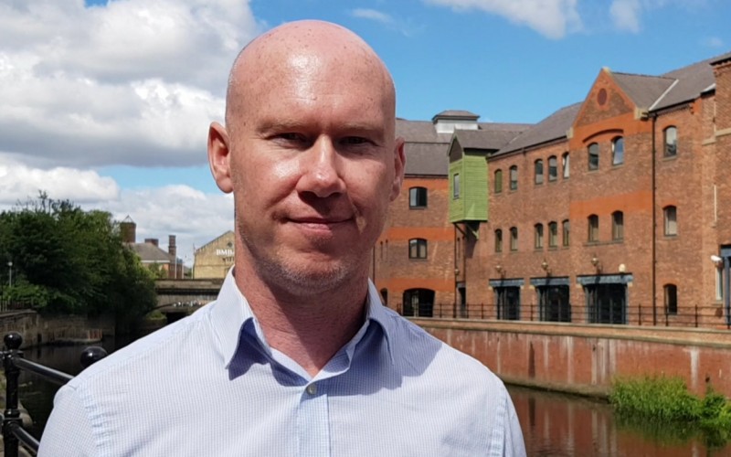 Michael Folan in open neck shirt standing in front of city centre river