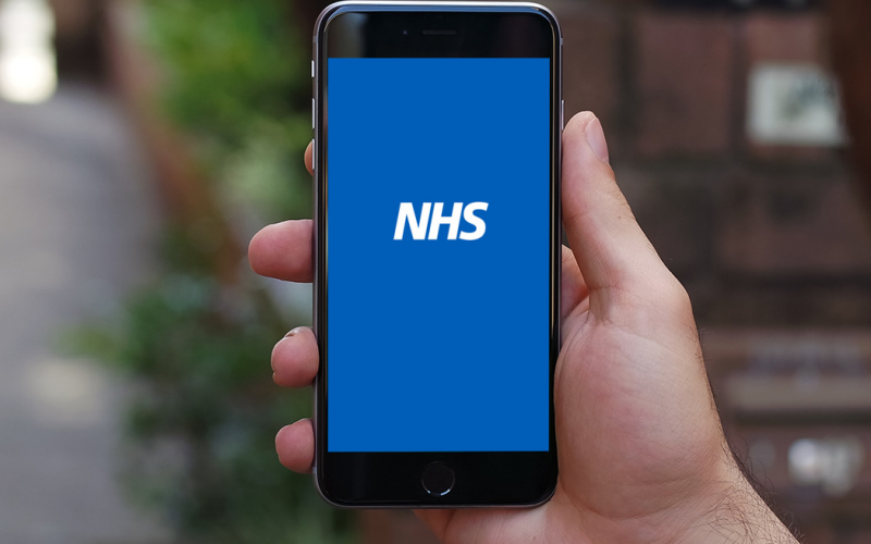 A hand holding a smart phone displaying an NHS logo on a plain blue background